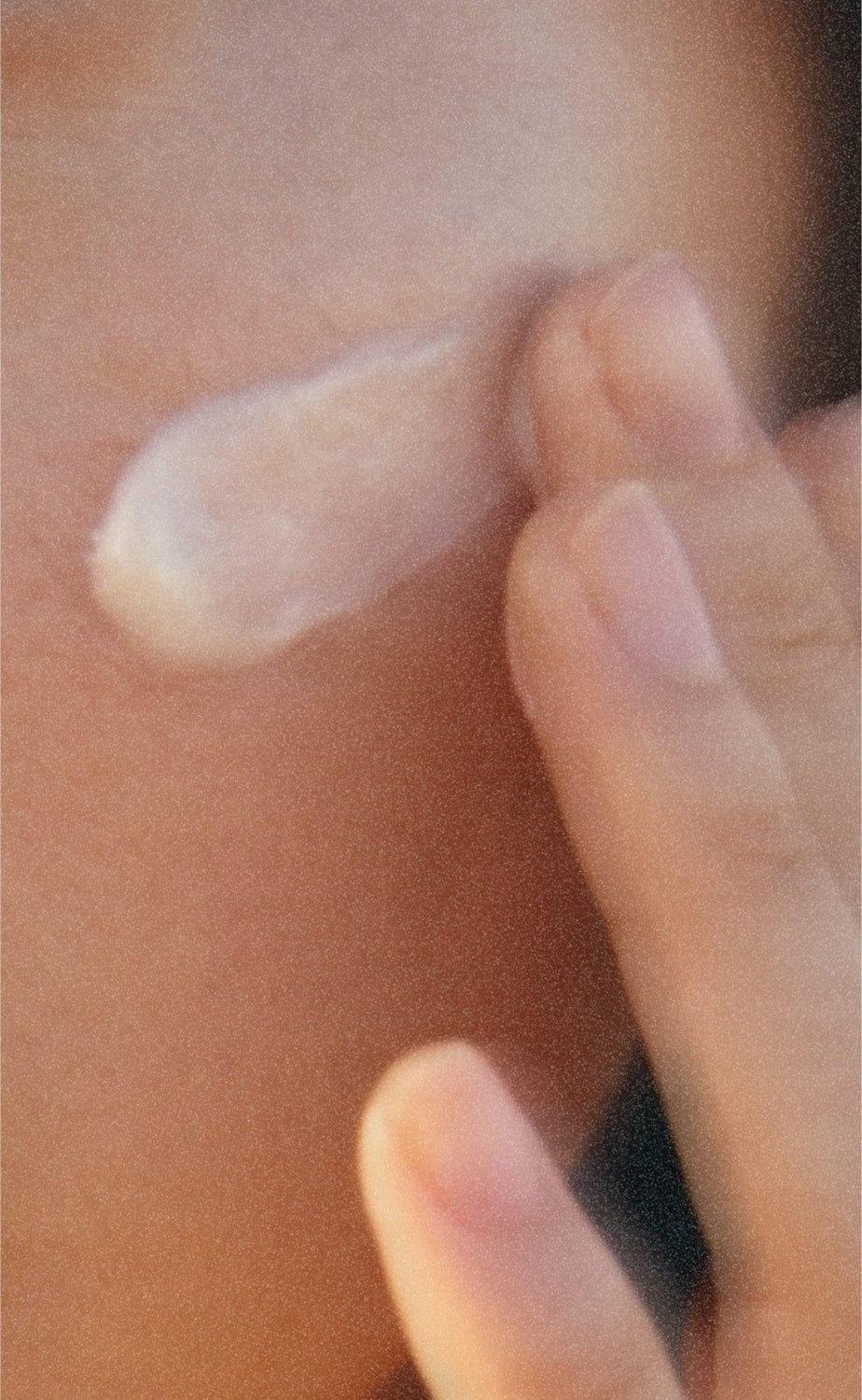 A fingertip presses cream into the cheek. Warm, unretouched skin.