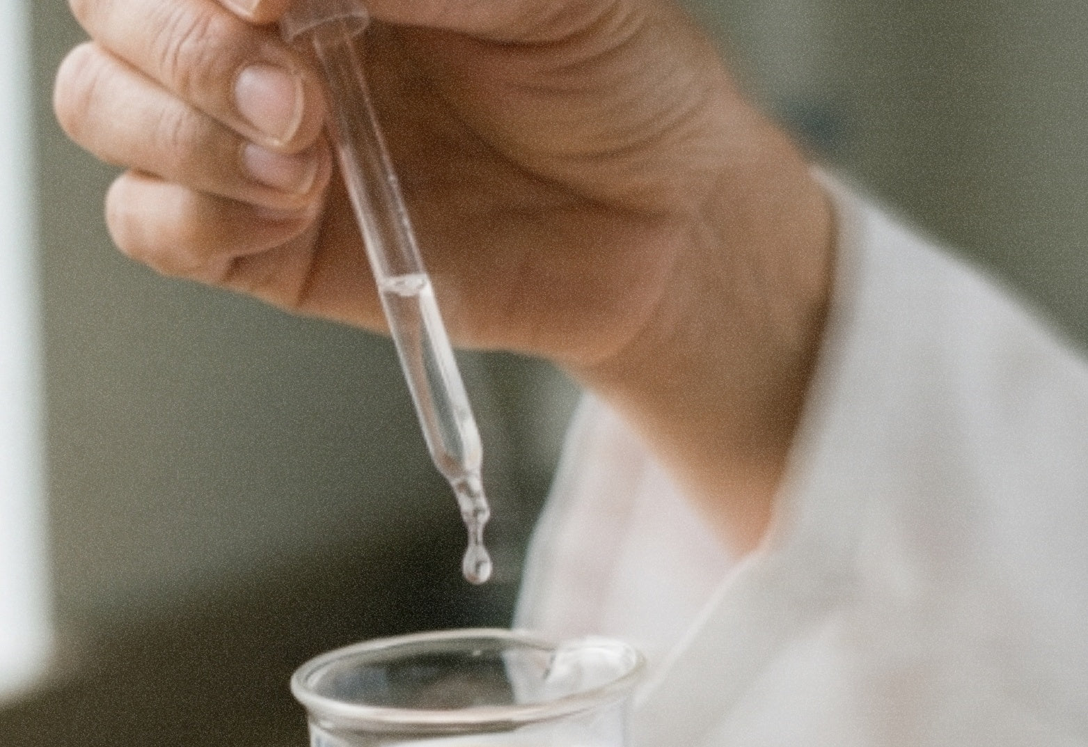 A formulator&rsquo;s hand pouring a dropper into a glass beaker.